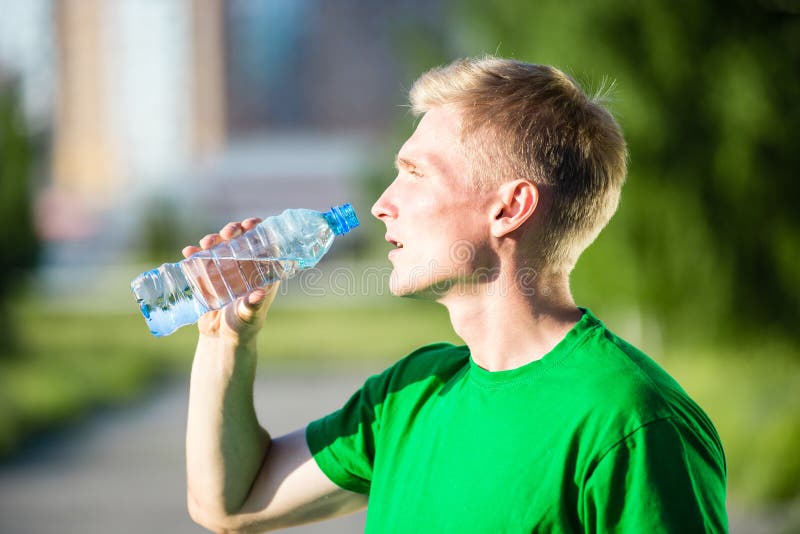 Tired man drinking water from a plastic bottle after fitness time and exercising in city street park at beautiful summer day. Sporty model caucasian ethnicity training outdoor. Tired man drinking water from a plastic bottle after fitness time and exercising in city street park at beautiful summer day. Sporty model caucasian ethnicity training outdoor.
