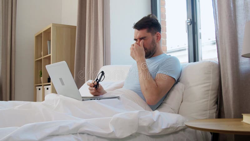 κουρασμένος άνδρας με γυαλιά με φορητό υπολογιστή στο κρεβάτι