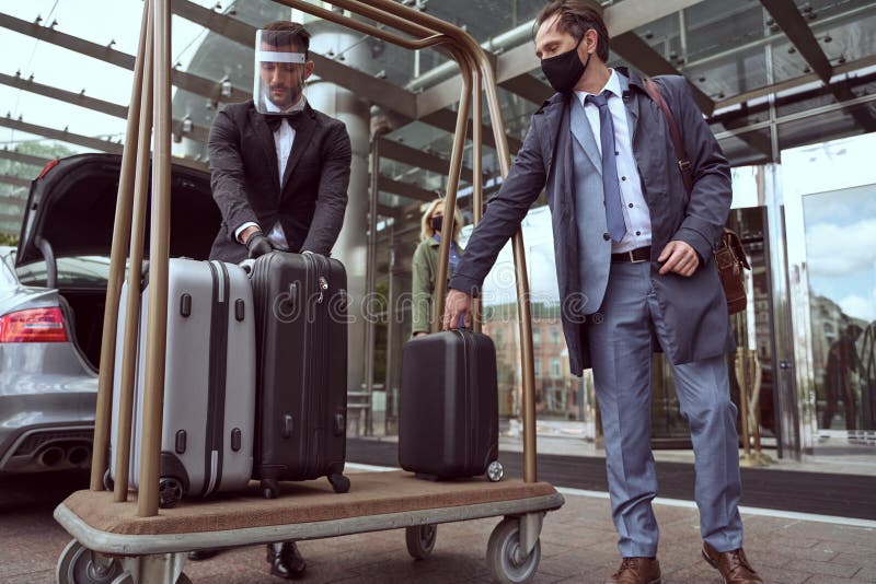 Weary-looking man in an elegant grey suit and protective mask getting his little black suitcase from a porters cart. Weary-looking man in an elegant grey suit and protective mask getting his little black suitcase from a porters cart