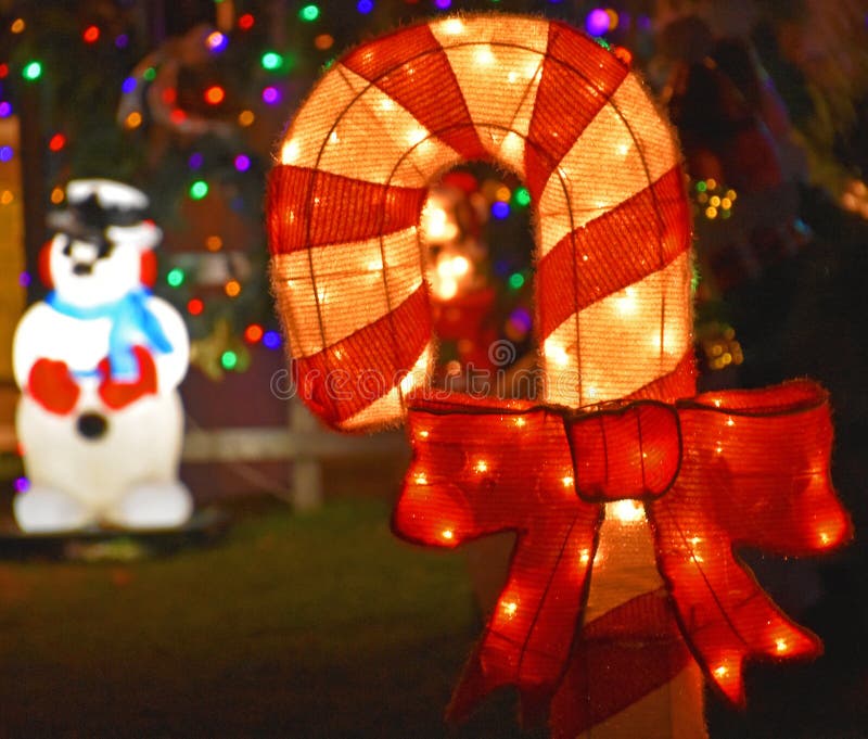 A large fabric candy cane lit with small lights inside with a red bow. Bokeh Christmas lights in the background, as well as a bright snowman with red gloves and blue scarf. A large fabric candy cane lit with small lights inside with a red bow. Bokeh Christmas lights in the background, as well as a bright snowman with red gloves and blue scarf.