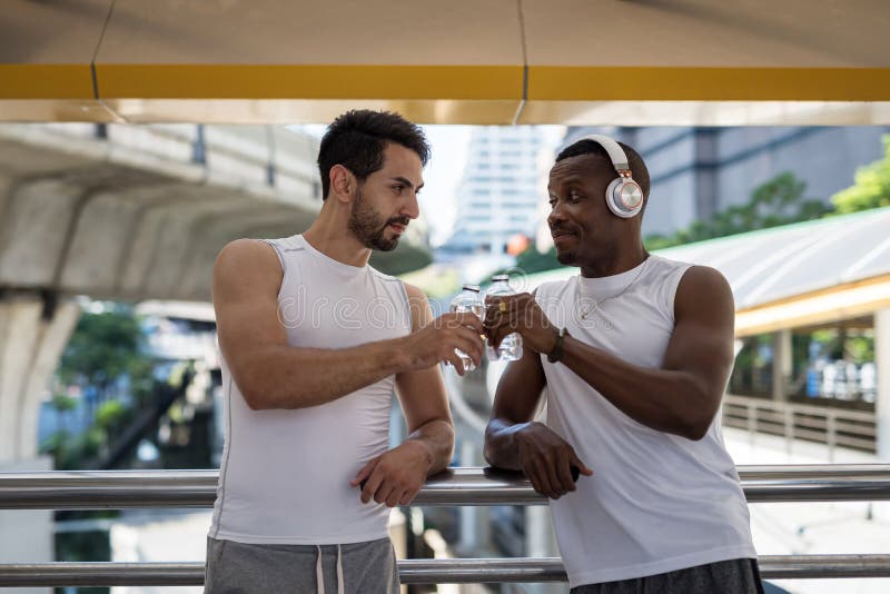 Happy American bearded and African black men toast and drink water after running workout in modern city. Bodybuilding and healthy lifetstyle. Lovely LGBT gay couple jogging exercise, active, adult, athlete, away, break, caucasian, clothing, day, fit, fitness, friends, handsome, happiness, laughing, lifestyle, look, male, man, mature, outdoors, park, people, relax, rest, smiling, sport, sweet, together, training, two, view, wellness. Happy American bearded and African black men toast and drink water after running workout in modern city. Bodybuilding and healthy lifetstyle. Lovely LGBT gay couple jogging exercise, active, adult, athlete, away, break, caucasian, clothing, day, fit, fitness, friends, handsome, happiness, laughing, lifestyle, look, male, man, mature, outdoors, park, people, relax, rest, smiling, sport, sweet, together, training, two, view, wellness