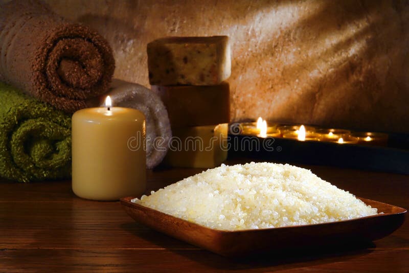 Natural bath sea salts in a wood bowl with cotton towels and artisan handmade soap bars gently lit by aromatherapy candles burning with a soft glow flame in a relaxation spa. Natural bath sea salts in a wood bowl with cotton towels and artisan handmade soap bars gently lit by aromatherapy candles burning with a soft glow flame in a relaxation spa