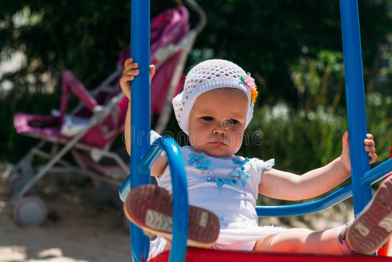 Λυπημένος, μικρό κορίτσι σε ένα άσπρο φόρεμα και καπέλο, που οδηγά σε μια ταλάντευση, έναν θερινό ήλιο και μια θερμότητα playgrou στοκ εικόνα με δικαίωμα ελεύθερης χρήσης Λυπημένος, μικρό κορίτσι σε ένα άσπρο φόρεμα και καπέλο, που οδηγά σε μια ταλάντευση, έναν θερινό ήλιο και μια θερμότητα playgrou στοκ εικόνα με δικαίωμα ελεύθερης χρήσης