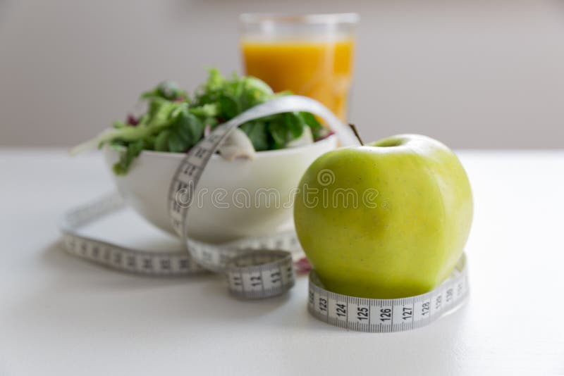 Measuring tape around the apple, bowl of green salad and glass of juice. Weight loss and right nutrition concept. Measuring tape around the apple, bowl of green salad and glass of juice. Weight loss and right nutrition concept.