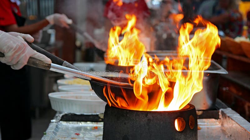 Professional chef in a commercial kitchen cooking flambe style. Chef frying food in flaming pan on gas hob in outdoor kitchen. Professional chef in a commercial kitchen cooking flambe style. Chef frying food in flaming pan on gas hob in outdoor kitchen.