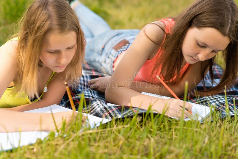 2 schoolgirls in the summer in the park are lying on a plaid, doing homework in a notebook. Concept of rest after school in nature. Emotion of concentration of study and knowledge. 2 schoolgirls in the summer in the park are lying on a plaid, doing homework in a notebook. Concept of rest after school in nature. Emotion of concentration of study and knowledge.