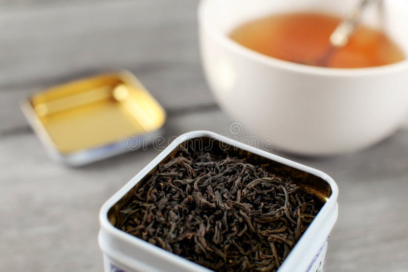 Black loose tea in metal tin, with blurred cup of hot amber drink in background. Black loose tea in metal tin, with blurred cup of hot amber drink in background