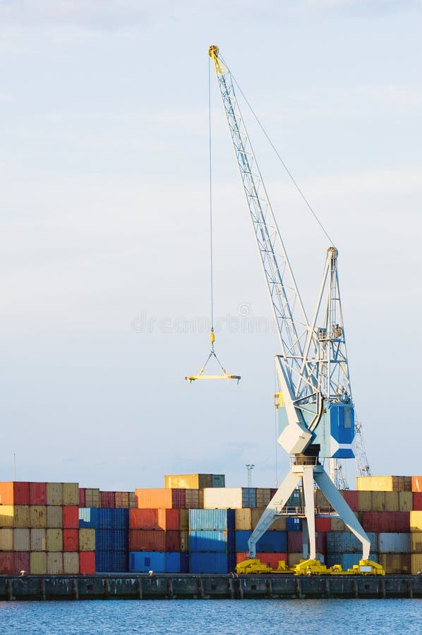 A large cargo crane stands at port in front of stacks of containers. There is no one viewable in the image. Vertically framed shot. A large cargo crane stands at port in front of stacks of containers. There is no one viewable in the image. Vertically framed shot.