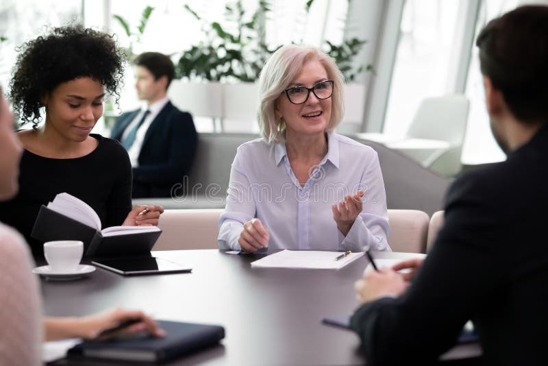 Aged female mentor conducting meeting with young professionals. Mix race business people group listen to experienced leader or coach while training. Young managers consulting with older businesswoman. Aged female mentor conducting meeting with young professionals. Mix race business people group listen to experienced leader or coach while training. Young managers consulting with older businesswoman