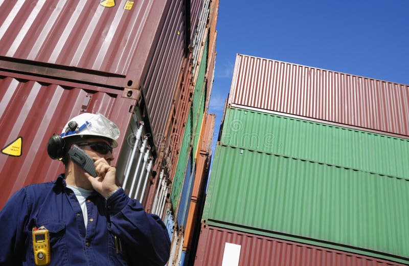 Dock and port worker talking in phone with stacks of freight containers in background. Dock and port worker talking in phone with stacks of freight containers in background