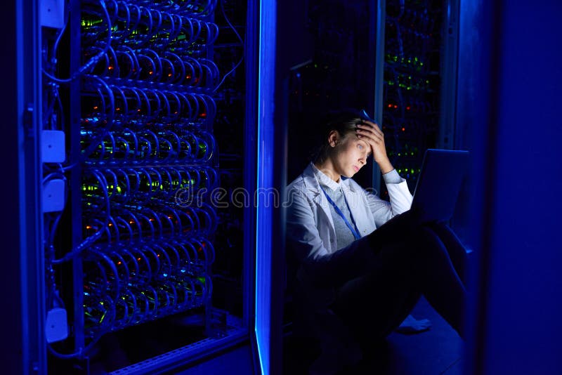 Portrait of tired young woman sitting on floor in dark server room using laptop while working with supercomputer at night. Portrait of tired young woman sitting on floor in dark server room using laptop while working with supercomputer at night