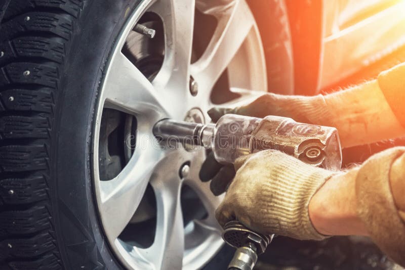 Car mechanic worker doing tire or wheel replacement with pneumatic wrench in garage of repair service station, sunlight effect, toned. Car mechanic worker doing tire or wheel replacement with pneumatic wrench in garage of repair service station, sunlight effect, toned
