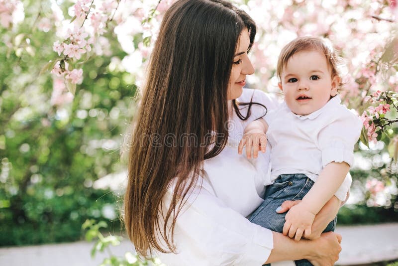 Μια ευτυχής μητέρα με ένα μωρό στα όπλα της στα άσπρα ενδύματα εξετάζει τα μάτια του στο κλίμα του άνθους κερασιών στοκ φωτογραφία Μια ευτυχής μητέρα με ένα μωρό στα όπλα της στα άσπρα ενδύματα εξετάζει τα μάτια του στο κλίμα του άνθους κερασιών στοκ φωτογραφία