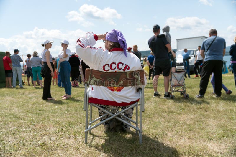 μια ηλικιωμένη γυναίκα με στολή με τη βοήθεια της επιγραφής στην πλάτη της κάθεται σε μια διπλωμένη καρέκλα στο γκαζόν και κοιτάζε