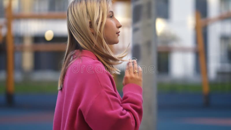 μια ξανθιά κοπέλα με ροζ πουλόβερ περπατάει σε ένα μοντέρνο συγκρότημα κατοικιών