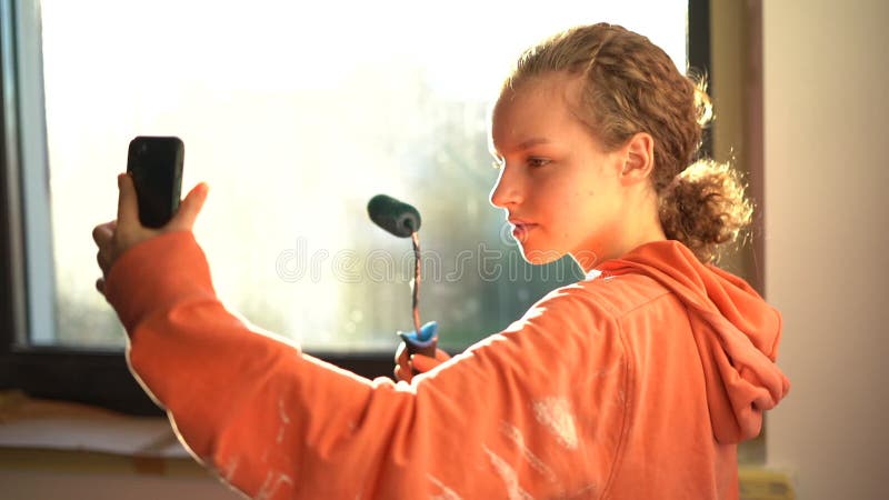 μια χαριτωμένη κοπέλα με κύλινδρο μπογιάς στα χέρια της παίρνει μια selfie ενώ ζωγραφίζει τους τοίχους στο δωμάτιό της. μέσα κοινω