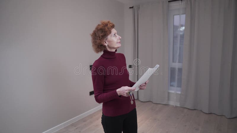 μια ώριμη γυναίκα εξετάζει ένα άδειο μικρό διαμέρισμα και έχει συνάψει συμφωνία ενοικίασης στο χέρι της. μισθώσεις ηλικιωμένων γυν απόθεμα βίντεο
