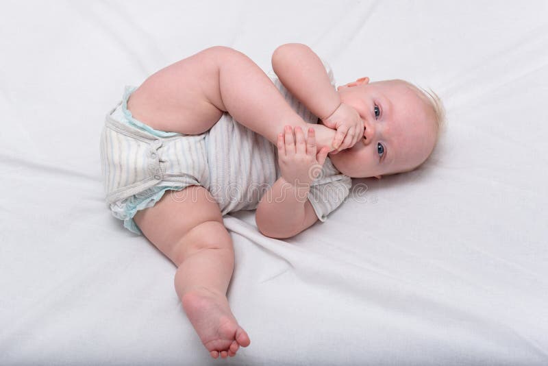 Little baby nibbles his foot on white bed. Cute 3 months baby. Gymnastics for the newborn. Little baby nibbles his foot on white bed. Cute 3 months baby. Gymnastics for the newborn.