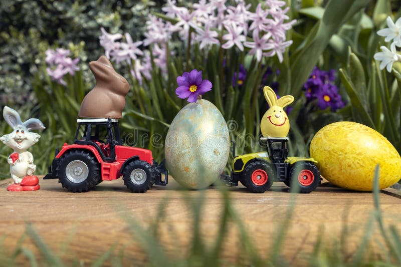 2 models of toy tractors, painted multi-colored eggs, chocolate and souvenir bunnies. Business concept for Easter greetings for farming. postcard. Grass in the foreground, flowers in the background. 2 models of toy tractors, painted multi-colored eggs, chocolate and souvenir bunnies. Business concept for Easter greetings for farming. postcard. Grass in the foreground, flowers in the background