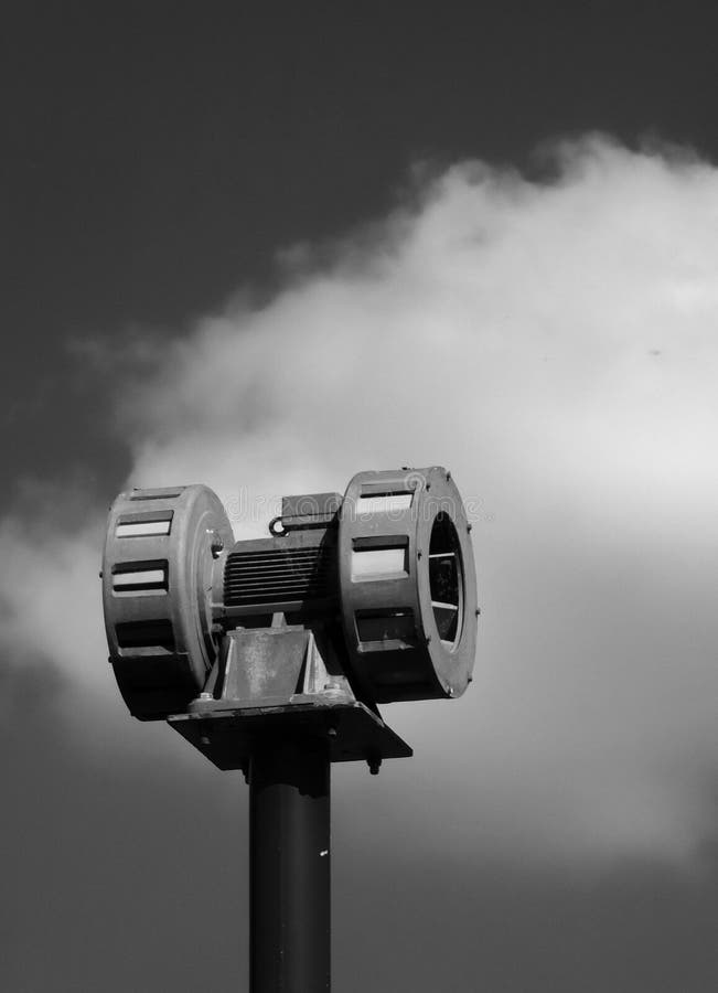 A monochrome image of an old mechanical warning siren on a pole against a cloudy sky. A monochrome image of an old mechanical warning siren on a pole against a cloudy sky