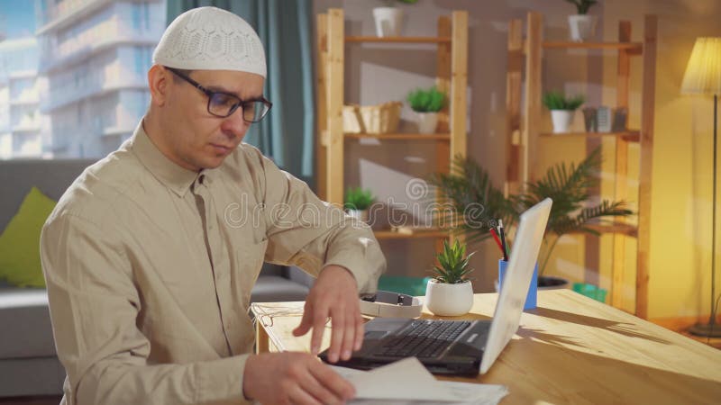 μουσουλμάνος με σκουφί εργάζεται στο σπίτι σε φορητό υπολογιστή