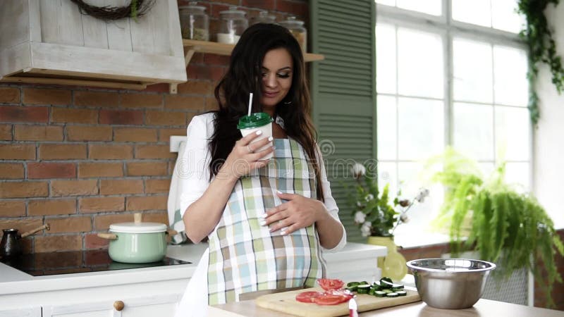 Μπροστινή άποψη της εγκύου γυναίκας σχετικά με την κοιλιά στην εσωτερική κουζίνα Καυκάσια γυναίκα στην ποδιά κουζινών που πίνει έ