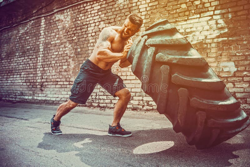 Muscular bearded tattooed fitness shirtless man moving large tire in street gym. Concept lifting, workout training. Muscular bearded tattooed fitness shirtless man moving large tire in street gym. Concept lifting, workout training.