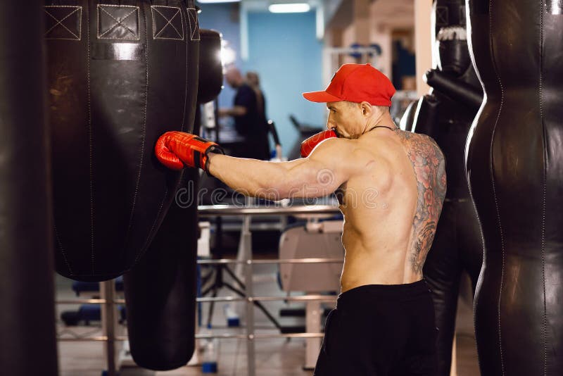 Μυϊκός μπόξερ γυμνοστήθων με punching την τσάντα στη γυμναστική Ένα άτομο με μια δερματοστιξία στα κόκκινα εγκιβωτίζοντας γάντια στοκ φωτογραφία Μυϊκός μπόξερ γυμνοστήθων με punching την τσάντα στη γυμναστική Ένα άτομο με μια δερματοστιξία στα κόκκινα εγκιβωτίζοντας γάντια στοκ φωτογραφία