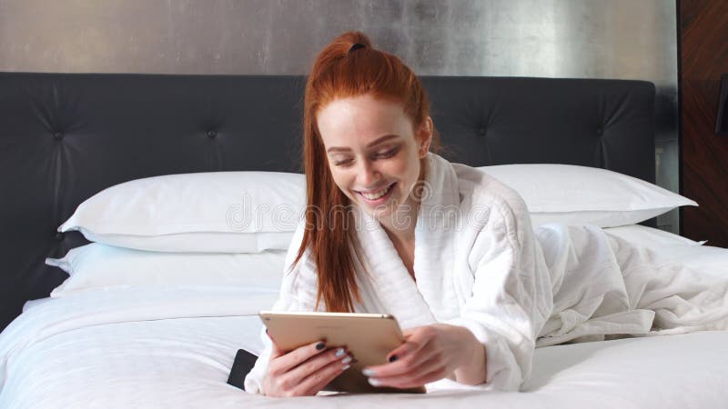 Νέα redhead γυναίκα που χρησιμοποιεί τον υπολογιστή ταμπλετών που βρίσκεται στο κρεβάτι στο δωμάτιο ξενοδοχείου