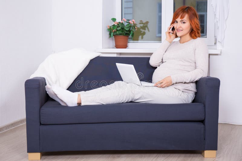 Νέα έγκυος γυναίκα χρησιμοποιώντας το lap-top και μιλώντας τηλεφωνικώς στο σπίτι στοκ εικόνες Νέα έγκυος γυναίκα χρησιμοποιώντας το lap-top και μιλώντας τηλεφωνικώς στο σπίτι στοκ εικόνες