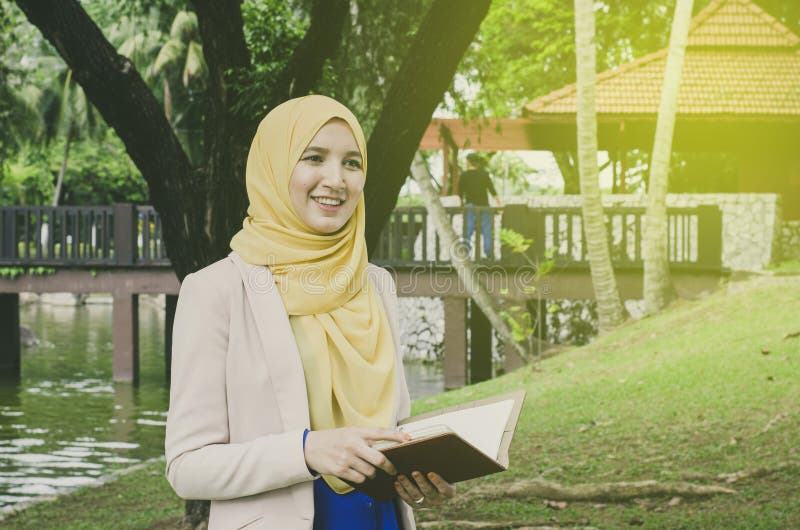 Νέα γυναίκα muslimah προσώπου χαμόγελου που στέκεται και που κρατά τα σημειωματάρια στο πάρκο Αρνητικό διάστημα για το κείμενο στοκ φωτογραφίες με δικαίωμα ελεύθερης χρήσης Νέα γυναίκα muslimah προσώπου χαμόγελου που στέκεται και που κρατά τα σημειωματάρια στο πάρκο Αρνητικό διάστημα για το κείμενο στοκ φωτογραφίες με δικαίωμα ελεύθερης χρήσης