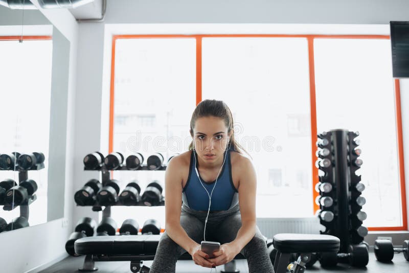 Young woman with earphones listening to music after hard workout in gym. Pretty sporty girl with ponytail is really tired and exhausted. She is looking at camera. Close up. Young woman with earphones listening to music after hard workout in gym. Pretty sporty girl with ponytail is really tired and exhausted. She is looking at camera. Close up