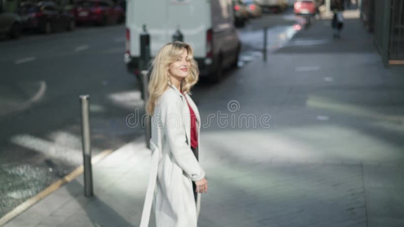 Νέα γυναίκα στο άσπρο παλτό που περπατά στην οδό και που κυματίζει στη κάμερα