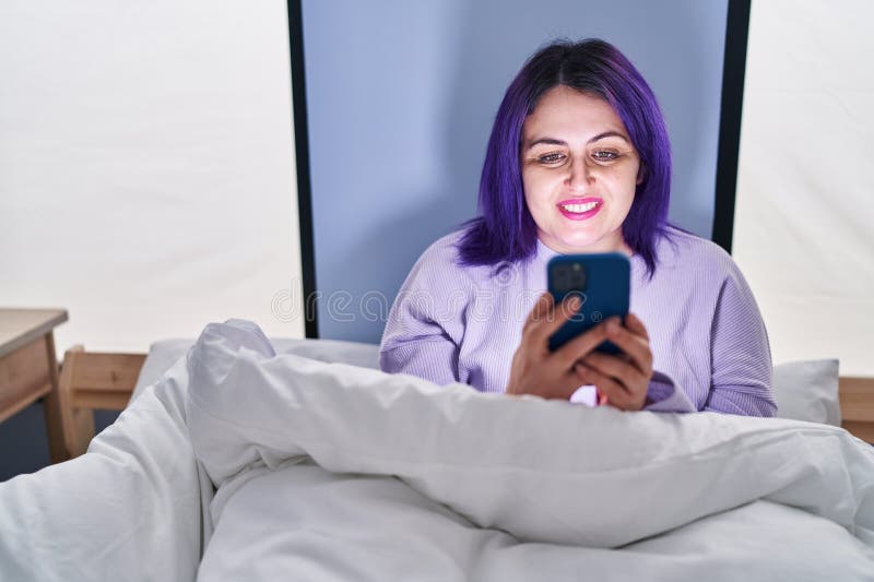 νέα όμορφη γυναίκα με μεγάλο μέγεθος, με smartphone να κάθεται στο κρεβάτι στοκ εικόνες
