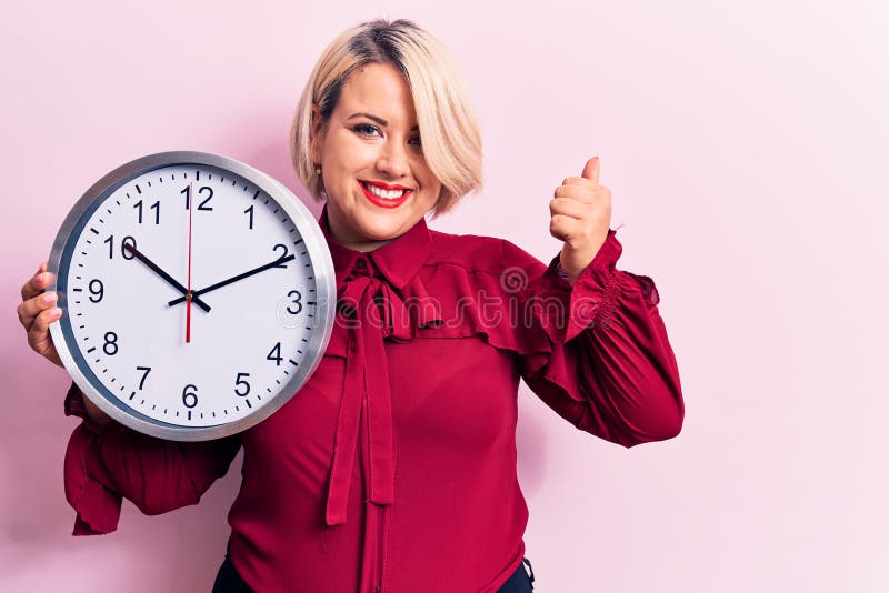 νέα όμορφη ξανθιά γυναίκα με μεγάλο μέγεθος που κάνει αντίστροφη μέτρηση χρησιμοποιώντας μεγάλο ρολόι πάνω από ροζ φόντο χαμογελών στοκ εικόνες