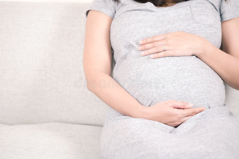 Νέα όμορφη συνεδρίαση εγκύων γυναικών στον γκρίζο καναπέ στο σπίτι και στοκ φωτογραφία με δικαίωμα ελεύθερης χρήσης Νέα όμορφη συνεδρίαση εγκύων γυναικών στον γκρίζο καναπέ στο σπίτι και στοκ φωτογραφία με δικαίωμα ελεύθερης χρήσης
