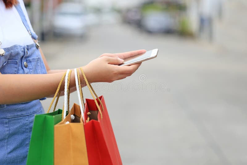 Νέες τσάντες smartphone και αγορών εκμετάλλευσης χεριών γυναικών με τη στάση στο χώρο στάθμευσης αυτοκινήτων στοκ εικόνα με δικαίωμα ελεύθερης χρήσης Νέες τσάντες smartphone και αγορών εκμετάλλευσης χεριών γυναικών με τη στάση στο χώρο στάθμευσης αυτοκινήτων στοκ εικόνα με δικαίωμα ελεύθερης χρήσης