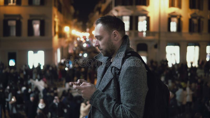 Νέο όμορφο άτομο τουριστών που στέκεται στο πλήθος το βράδυ Η αρσενική χρήση το smartphone βρίσκει την κατεύθυνση