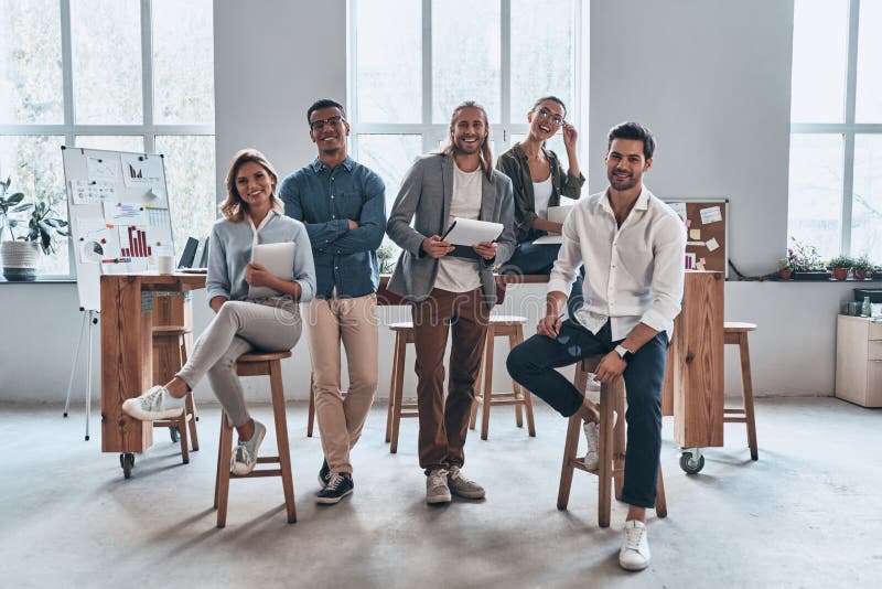 Young business professionals. Full length of young modern people in smart casual wear smiling and looking at camera while standing in the creative office. Young business professionals. Full length of young modern people in smart casual wear smiling and looking at camera while standing in the creative office