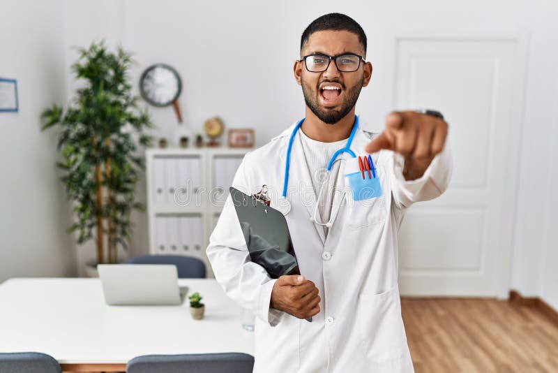 Young indian man wearing doctor uniform and stethoscope pointing displeased and frustrated to the camera, angry and furious with you. Young indian man wearing doctor uniform and stethoscope pointing displeased and frustrated to the camera, angry and furious with you