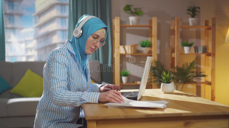 νέος μουσουλμάνος ελεύθερος επαγγελματίας σε εθνική μαντίλα εργάζεται σε φορητό υπολογιστή που κάθεται στο σαλόνι