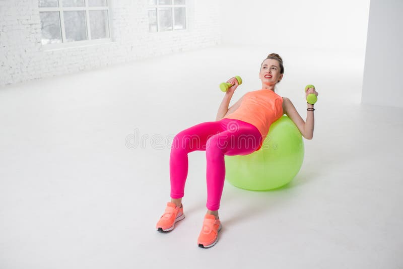 Tired woman in colorful sportswear pushing dumbbels lying on the fitness ball in the white gym. Tired woman in colorful sportswear pushing dumbbels lying on the fitness ball in the white gym