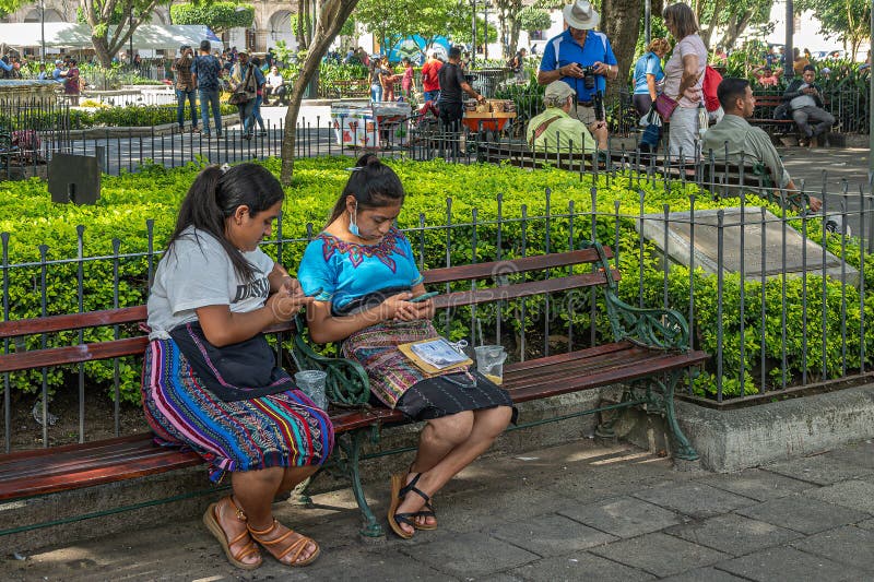 Guatemala, La Antigua - July 20, 2023: 2 young women, in colorful traditional clothing, read on their smartphones sitting on bench in park of Plaza Mayor, central square. Guatemala, La Antigua - July 20, 2023: 2 young women, in colorful traditional clothing, read on their smartphones sitting on bench in park of Plaza Mayor, central square