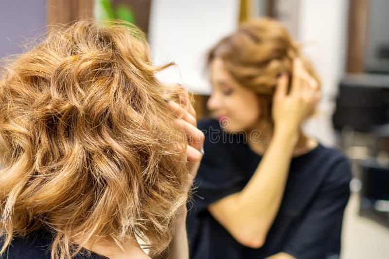 νεαρή γυναίκα που κοιτούσε το νέο σγουρό καστανό στυλ της μπροστά στον καθρέφτη στο κομμωτήριο στοκ φωτογραφίες