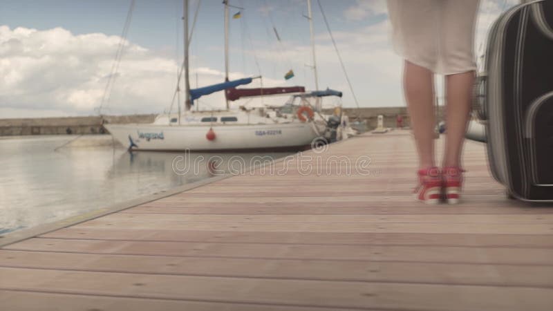 νεαρή γυναίκα που πηγαίνει διακοπές. γυναίκα με βαριά βαλίτσα. θηλυκό που περπατά μακριά στην ξύλινη γέφυρα στο λιμάνι του γιοτ απόθεμα βίντεο