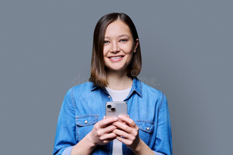 Young happy woman using smartphone in gray background. Smiling 20s female looking at camera texting. Mobile Internet apps applications technologies for work education communication shopping healthcare. Young happy woman using smartphone in gray background. Smiling 20s female looking at camera texting. Mobile Internet apps applications technologies for work education communication shopping healthcare