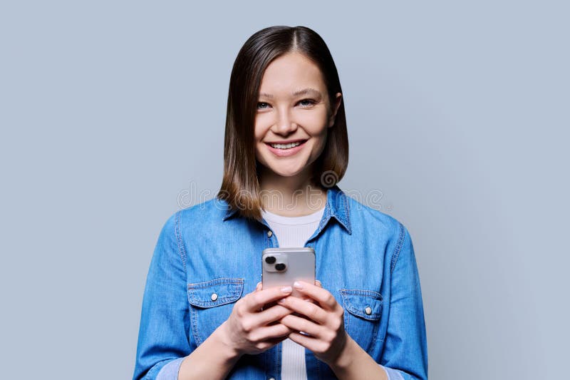 Young happy woman using smartphone in gray background. Smiling 20s female looking at camera texting. Mobile Internet apps applications technologies for work education communication shopping healthcare. Young happy woman using smartphone in gray background. Smiling 20s female looking at camera texting. Mobile Internet apps applications technologies for work education communication shopping healthcare