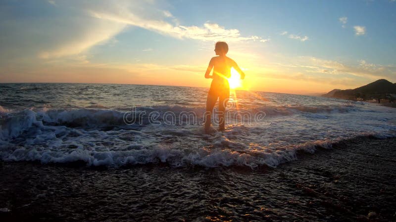 νεαρή γυναίκα τρέχει και σηκώνει τα χέρια ψηλά στο χρυσό ηλιοβασίλεμα στην παραλία σε κύματα