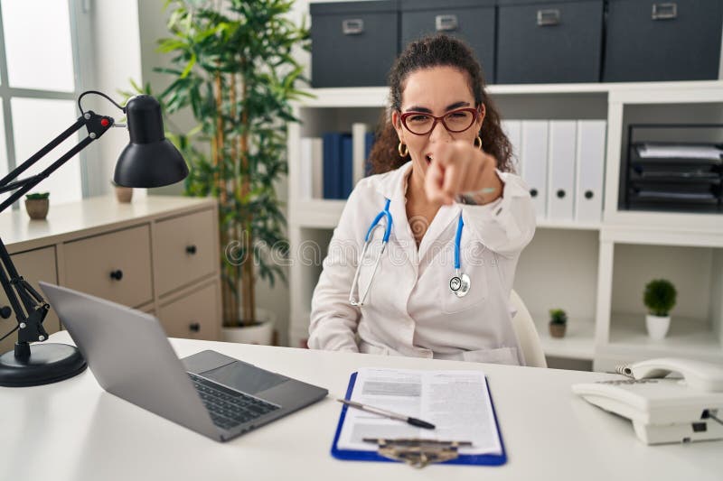 Young hispanic woman wearing doctor uniform and stethoscope pointing displeased and frustrated to the camera, angry and furious with you. Young hispanic woman wearing doctor uniform and stethoscope pointing displeased and frustrated to the camera, angry and furious with you