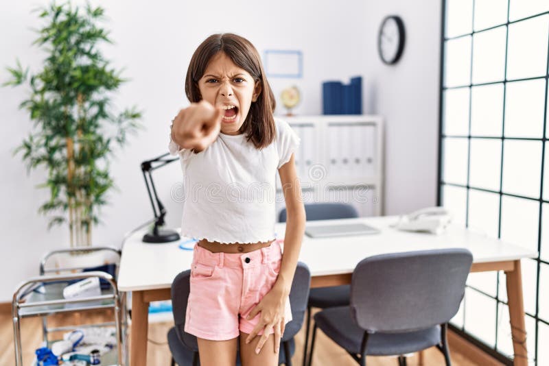 Young hispanic girl standing at pediatrician clinic pointing displeased and frustrated to the camera, angry and furious with you. Young hispanic girl standing at pediatrician clinic pointing displeased and frustrated to the camera, angry and furious with you